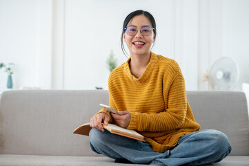 Obraz premium A happy Asian woman sitting on a couch with a book in her hand and smiling at the camera.