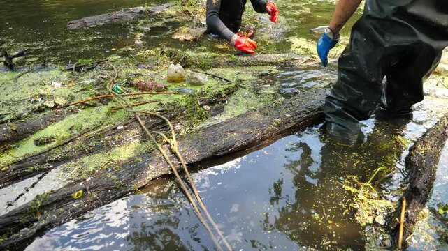 Unrecognizable male eco activists in rubber suits collecting debris in dirty lake. Volunteers in protective gloves cleaning messy river of rubbish. Concept of environment pollution protection. Slow mo