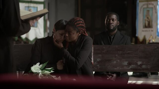 African American woman consoling Caucasian female friend touching her face and wiping tears during sermon at funeral ceremony in church, Black man sitting in background