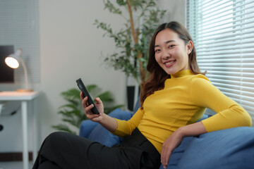 Happy attractive Asian woman holding mobile phone using technology for online shopping, e-commerce, buying things online, texting, greeting, chatting, relaxing on sofa in living room at home.