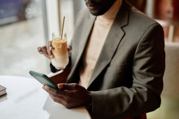 Man holding smartphone while sipping latte in a cozy cafe interior. Window allows natural light to illuminate his workspace and beverage while he engages on his phone