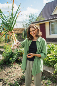 Happy woman holding harvested garlic and standing at farm near house