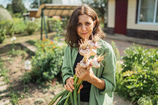 Smiling woman holding fresh garlic and standing at farm near house