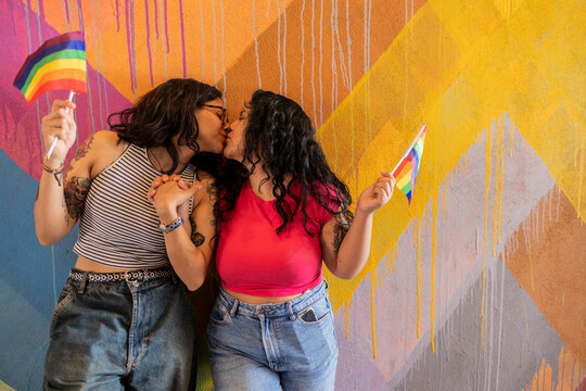 Affectionate lesbian couple holding pride flag and kissing near painted wall
