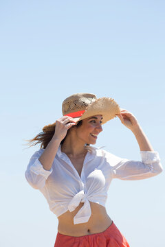 Happy woman wearing knotted shirt holding hat and standing under clear sky