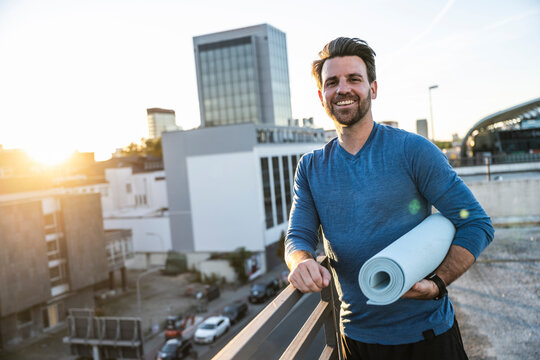 Happy man holding rolled up exercise mat leaning on railing at terrace
