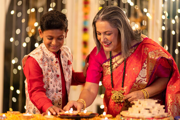 Happy grandmother with grandson lighting diyas while sitting on chair in illuminated home during...