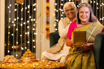 Happy senior husband giving surprise gift to excited wife in illuminated home on occasion of Diwali...