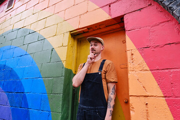 Playful gay person standing with finger on lips in front of colored door