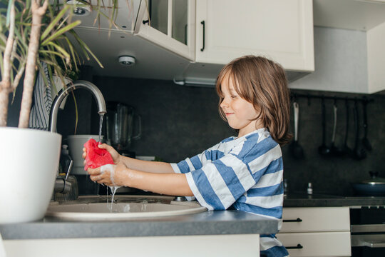Cute boy holding cleaning sponge under running water in kitchen