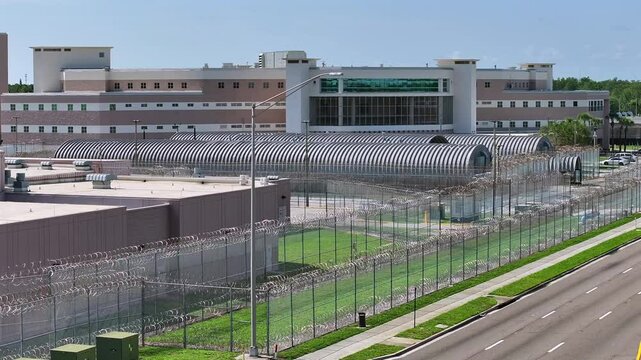 A large detention facility surrounded by barbed wire fences and security infrastructure in Florida, with multiple levels and fortified perimeter areas. Roads run parallel to the facility. Aerial.