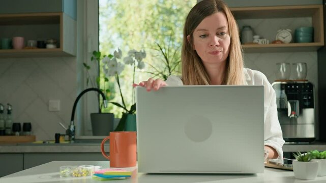 Female freelancer wraps up her working day by closing laptop in kitchen. Woman finishes her work at desk in home office with laptop and stationery