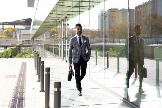 Businessman with hand in pocket walking on sidewalk outside glass building