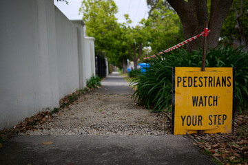 Bright yellow pedestrians watch your step sign, with black text, near a piece of unpaved footpath, in a leafy green suburb during an overcast day