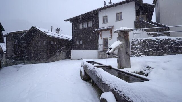 Snow Falling At Water Trough And Alpine Houses During Winter In Blatten bei Naters Village In Valais, Switzerland. - static shot