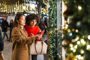 Two young women window shopping while checking smartphone in a decorated city at christmas time