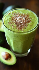 A tall glass of green smoothie topped with shredded coconut and flax seeds. There are two avocado halves in the background.