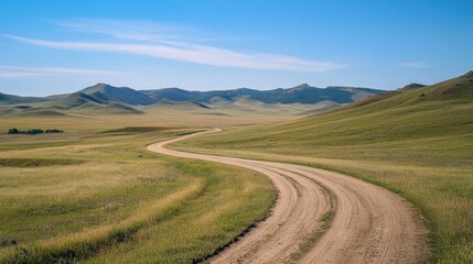 Fototapeta premium A winding dirt road stretches across open grasslands with mountains in the background and a clear blue sky overhead