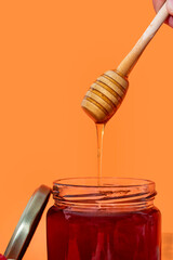 Glass jar full of honey and wooden dipper with honey drop isolated on orange background