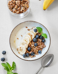 Flat lay of granola with yogurt, banana and blueberries in a bowl on a light background with spoon and mint.