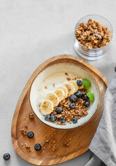 Natural yogurt with granola or muesli, banana and blueberries in a bowl on a wooden board on a light background