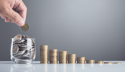 Hand drops coins into a glass jar, with stacks of coins arranged from smallest to largest on the...