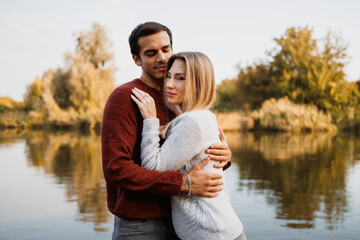 Man in sweater hugging girlfriend near blurred lake in autumn