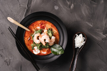 Spicy soup with shrimp, mushrooms, lemon, coconut milk and chili pepper in a black bowl on a dark table background. Tom Yum Kung with chopsticks and a spoon with boiled rice. Thai food