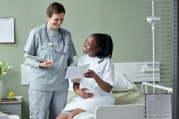 Obraz premium Doctor engaging with smiling child patient sitting on hospital bed holding papers in bright medical room with equipment visible around