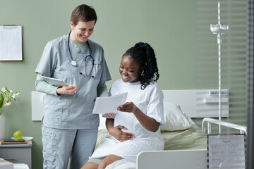 Nurse engaging with patient in hospital room, both smiling while reviewing documents Nurse standing next to bed, patient sitting up