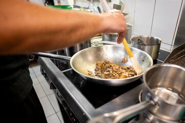 person cooking in a kitchen, focusing on a frying pan with sautéed mushrooms over a stove. Visible steam suggests the food is hot. The scene captures the action of cooking. Ideal for culinary themes.