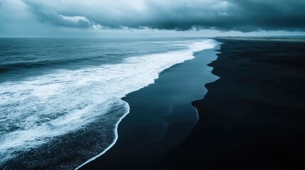 Fototapeta premium Dark black sand stretches across the beach as the deep blue ocean waves gently roll in under a cloudy sky.