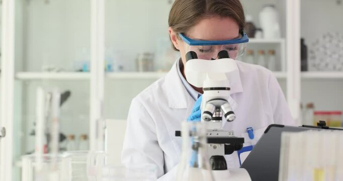Woman biochemist with protective goggles analyzes samples under microscope in laboratory setting. Focused chemist researches material in lab