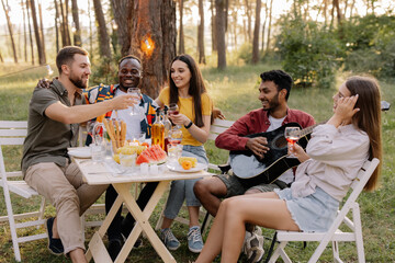 Meeting of multiracial group of friends playing guitar, singing, eating dinner and drinking wine during party in the forest