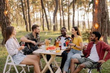 Meeting of multiracial group of friends playing guitar, singing, eating dinner and drinking wine during party in the forest