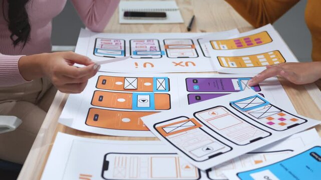 Two user experience designers collaborate at a desk, arranging cards to create a prototype for a mobile application's user interface, focusing on usability and user flow