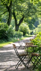 A row of empty cafe tables and chairs sit on a gravel path in a park setting, with lush greenery in the background.