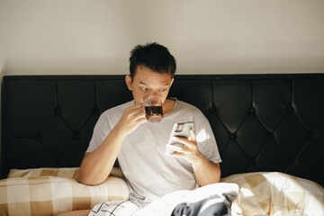 Asian man enjoy drinking coffee while holding smartphone on the bed