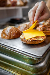 A close-up of a hand placing a slice of cheese on an open sandwich, with more sandwiches in the background. The scene is set on a wooden table, highlighting the fresh ingredients and homemade feel.
