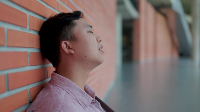 Close-up of Asian man's face sitting with a very disappointed and sad expression, leaning against a brick wall, kneeling, hands on knees and head on his head. He feels sad, contemplative, lost and dis