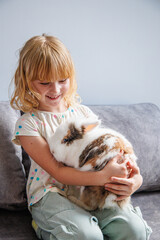 A young girl joyfully cuddling a fluffy rabbit while sitting on a gray couch indoors