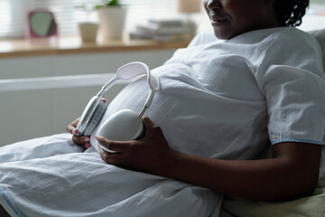 Pregnant woman sitting comfortably in living room, holding prenatal monitor for listening to baby's heartbeat. Room adorned with soft furniture and ambient light