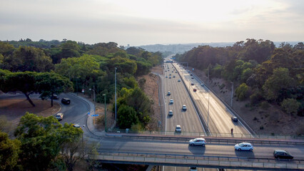 Autoestrada em Monsanto, Lisboa, ao pôr do sol com tráfego em movimento. O céu alaranjado ilumina a estrada e os veículos, criando um contraste entre a natureza e a paisagem urbana.