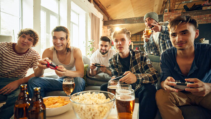 Living room alive with energy as guys laugh and focus on action, controllers in hand. Popcorn, chips, and beers complete ultimate game session. Concept of football fans, sport evens, cybersport.