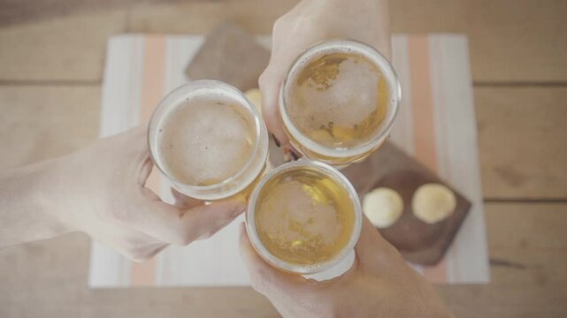 Overhead view of three people toasting with glasses of beer and enjoying traditional chipa bread on a wooden table with a striped tablecloth