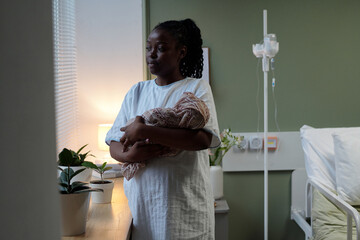 Mother standing by hospital room window holding newborn baby wrapped in blanket gazing...