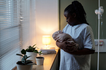 African American mother holding newborn baby while standing by window in hospital room with potted...
