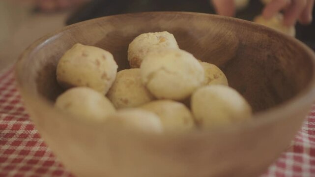 Close up footage shows a person person putting freshly baked chipa bread buns in a rustic wooden bowl, placed on a red and white checkered cloth, capturing the essence of homemade traditional cuisine