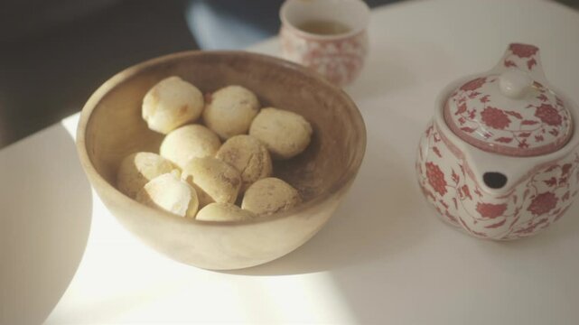 Sunlight illuminates womans hand grabbing freshly baked chipa bread from wooden bowl, accompanied by a floral teapot and cup on a white table, creating a cozy home atmosphere footage