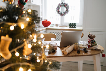 Cozy kitchen scene featuring Christmas decorations, laptop on wooden table, warm lights from Christmas tree. Festive atmosphere with baking ingredients and holiday mood.